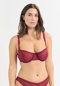 Burgundowa koronkowa biustonosz z fiszbinami, skomplikowany wzór geometryczny, falbany na brzegach, regulowane ramiączka oraz mały dekoracyjny detal.