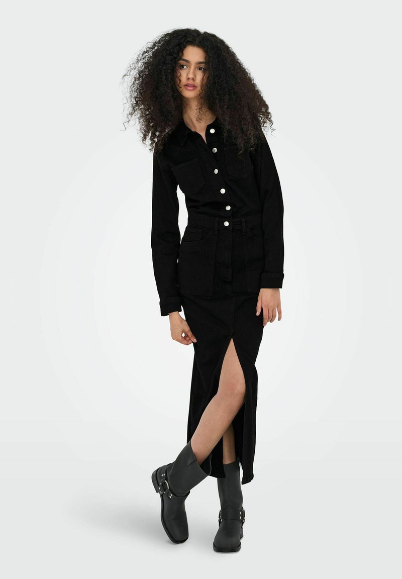 Femme aux cheveux bouclés portant une chemise noire boutonnée, une jupe noire fendue haute et des bottines noires, debout avec une jambe croisée.