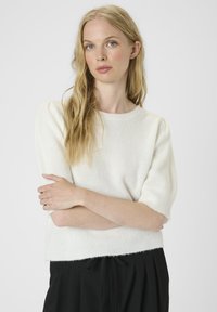 Blonde vrouw met golvend haar, gekleed in een wit kortemouwen truitje en zwarte broek, staand met gekruiste armen tegen een effen achtergrond.