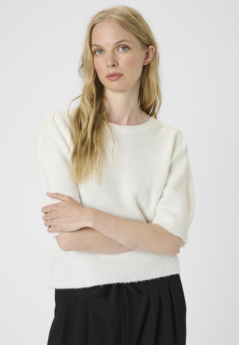 Blonde vrouw met golvend haar, gekleed in een wit kortemouwen truitje en zwarte broek, staand met gekruiste armen tegen een effen achtergrond.