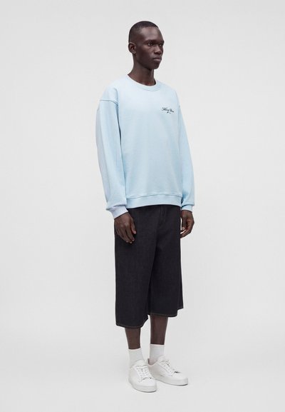 Homme portant un sweat-shirt bleu clair, un short large noir jusqu’aux genoux, des chaussettes blanches et des baskets blanches, debout devant un fond uni.