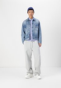 Leren spijkerjack, lichtblauw, met twee voorzakken, gedragen over een lichtpaarse overhemd met knopen. Grijze joggingbroek en witte sneakers.
