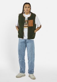 Colete de polar verde com bolso castanho, gola redonda e fecho éclair. Usado sobre uma t-shirt branca gráfica e combinado com jeans azul claro e ténis.