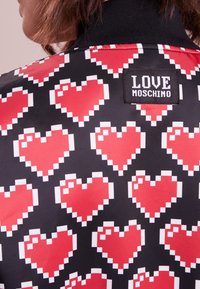 Black jacket featuring a repeated pixelated heart pattern in red and white. Includes a black label with "LOVE MOSCHINO" branding.