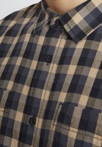 Camicia a bottoni con un motivo a quadri nero e tan; presenta un collo piatto, bottoni neri e una tasca sul petto. Realizzata in tessuto testurizzato.