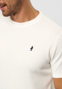 Hvid, ribbet t-shirt med korte ærmer, der har en lille marineblå, broderet figur på brystet. Rund halsudskæring.