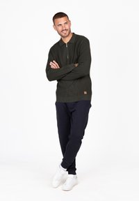 Dunkelgrüner Pullover mit Reißverschlusskragen, strukturiertem Stoff und kleinem braunem Logo-Patch, kombiniert mit navyblauen Hosen und weißen Sneakers.