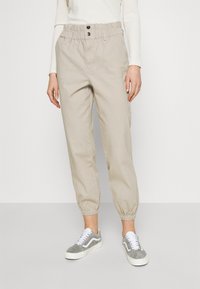 Personne portant un pantalon fuselé beige à taille élastique et des baskets gris-blanc, debout devant un fond clair uni.