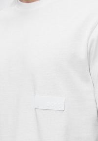T-shirt blanc à col rond avec un petit logo blanc en patch indiquant "BOSS" sur le bas du côté droit de la poitrine.