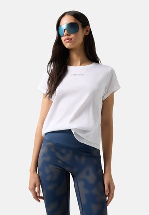 Kobieta o długich ciemnych włosach, nosząca niebieskie lustrzane okulary przeciwsłoneczne, białą koszulkę z napisem "FIRE+ICE" oraz niebieskie legginsy w wzory, stojąca na jednolitym tle.