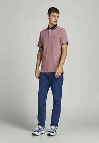 Jovem de pé, vestindo uma camisa polo rosa com colarinho e punhos listrados, jeans azuis e ténis brancos, contra um fundo claro e neutro.