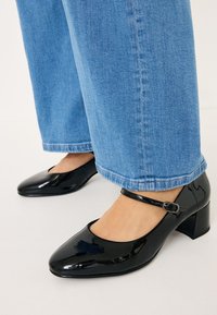 Black patent leather heels with a square toe, ankle strap, and a block heel, paired with light blue wide-leg jeans.