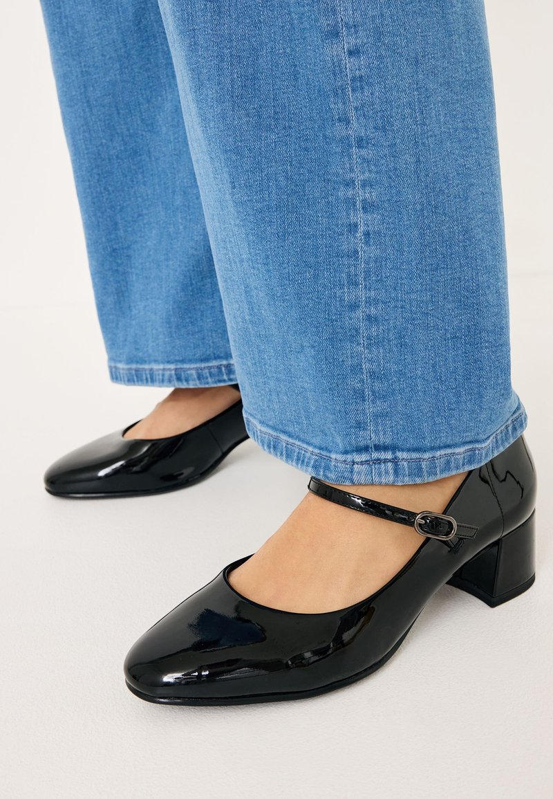 Black patent leather heels with a square toe, ankle strap, and a block heel, paired with light blue wide-leg jeans.