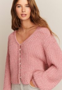 Lyserød, fluffy cardigan med V-hals, ribbet struktur, frontlukning med knapper med sølvdetaljer og løse, lange ærmer.