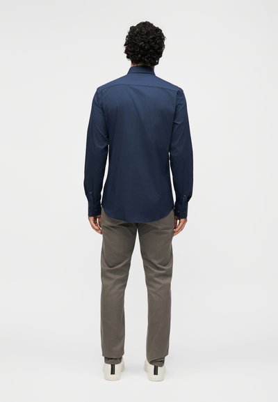 Άνδρας με σγουρά μαλλιά που φοράει μπλε σκούρο μπλουζάκι μακρυμάνικο, γκρι παντελόνι και λευκά αθλητικά παπούτσια, στέκεται πλάτη σε ένα λευκό φόντο.