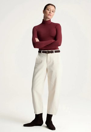 Kobieta stoi z rękami skrzyżowanymi na piersi, ubrana w bordowy golf, białe spodnie z wysokim stanem i paskiem oraz czarne botki na tle jednolitego tła.