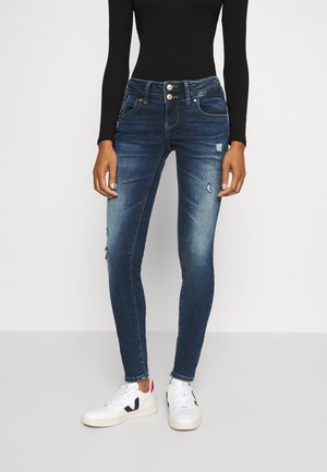 Femme portant un jean skinny bleu foncé usé avec une taille à double bouton et des baskets blanches, debout devant un fond uni.