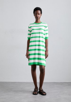 Gestreepte katoenen t-shirt jurk in groen en wit, met korte mouwen en een ontspannen pasvorm, tot halverwege de dijen reikend. Gdragen met zwarte sandalen.
