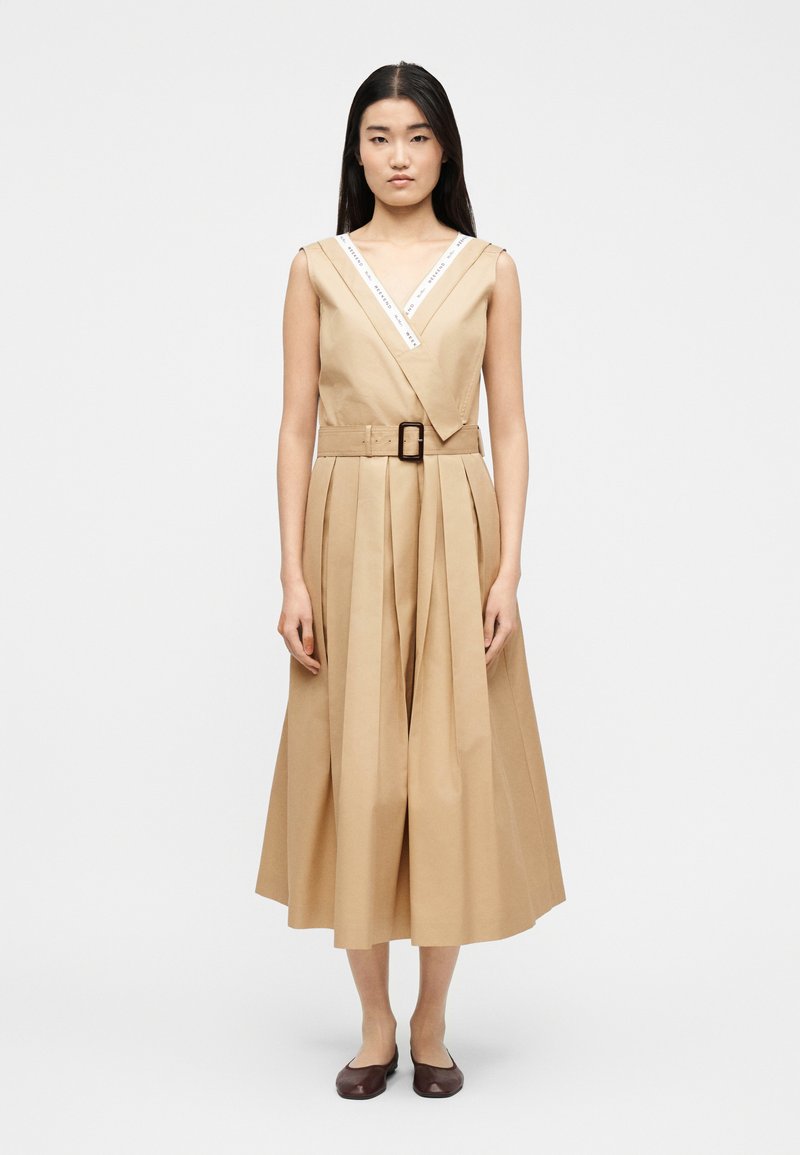 Γυναίκα όρθια που φοράει αμάνικο μπεζ midi φόρεμα με πλισέ φούστα, ντεκολτέ σε σχήμα V με λευκή διακοσμητική ρίγα, ζώνη και σκούρα επίπεδα παπούτσια σε λευκό φόντο.