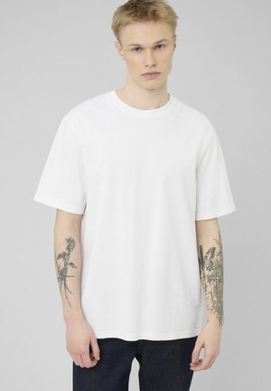 Jeune homme aux cheveux blonds courts portant un T-shirt blanc uni et un pantalon foncé, montrant des tatouages sur les deux avant-bras sur un fond gris.