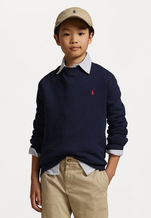Maglione blu navy con logo rosso, indossato sopra una camicia a righe leggera blu chiaro con colletto. Pantaloni beige casual e un cappellino beige completano l'outfit.