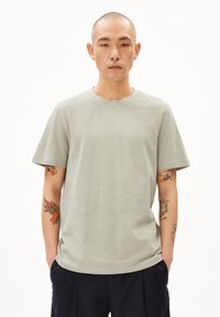Hellgrünes T-Shirt mit kurzen Ärmeln und Rundhalsausschnitt, strukturiertem Stoff, lockerer Passform und sichtbaren Tattoos an den Armen des Models.