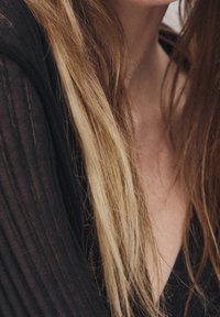 Schwarzer, strukturierter Stoff fällt über die Schulter und enthüllt einen tiefen V-Ausschnitt. Langes Haar mit helleren Strähnen fließt sanft herab.