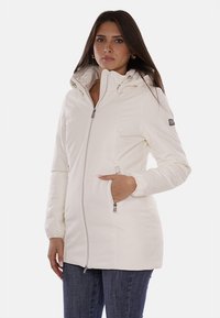 Giacca isolante bianca con colletto alto e chiusura a zip. Presenta polsini elasticizzati, due tasche laterali e un patch con logo sulla manica.