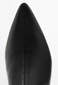 Scarpa nera in pelle con punta appuntita caratterizzata da una texture liscia e cuciture visibili lungo il lato, evidenziando il suo design elegante.