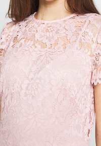 Detailní záběr na světle růžový krajkový top s květinovými vzory, krátkými rukávy a kulatým výstřihem, který má na sobě osoba.