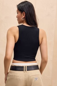 Frau mit langen dunklen Haaren trägt ein schwarzes ärmelloses Croptop, beige Hose mit schwarzem Gürtel und silbernen Creolen und schaut von der Kamera weg.