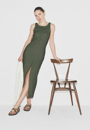 Kvinde i olivengrøn ærmeløs kjole med frontslids læner sig op ad en træstol, holder et hvidt tekstureret stof og står foran en ensfarvet hvid baggrund.