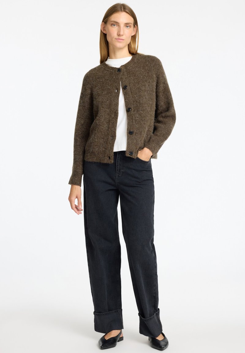 Braune Strickjacke mit Rundhalsausschnitt und Knöpfen vorne, kombiniert mit einem weißen Oberteil und schwarzen weiten Jeans. Schlichtes, texturiertes Design.