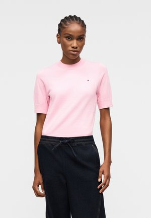 Kobieta z warkoczami, ubrana w jasno różowy krótki rękaw i czarne spodnie ze ściągaczem, stoi na prostym jasnym tle.