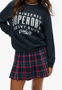 Sudadera azul marino con gran texto blanco, combinada con una falda de cuadros plisada que presenta colores rojo, verde y azul marino. Ambos son atuendos casuales.