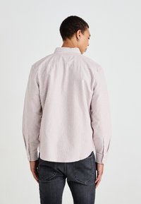 Striped button-up shirt in light pink and white cotton, featuring a collar, long sleeves, and a curved hem. Worn with dark jeans.