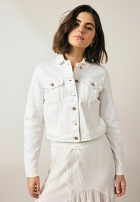 Chaqueta de denim blanca con un diseño corto, dos bolsillos en el pecho y cierres de botón plateados. La textura es suave con un detalle de costura mínimo.