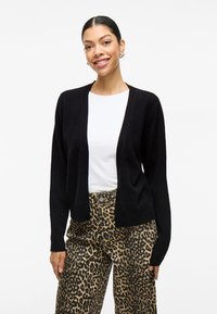 Μαύρο καρντιγκάν με ανοιχτό μπροστινό σχέδιο, ριμπ μανσέτες και μαλακή υφή, φορεμένο πάνω από λευκό πουκάμισο και συνδυασμένο με παντελόνι με λεοπάρ σχέδιο.