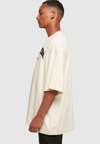 Beige oversized t-shirt med korte ærmer, med sort trykt tekst på forsiden. Blødt bomuldsstof, afslappet pasform, detalje i sidesøm.