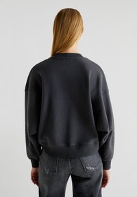 Felpa oversized grigio scuro con collo a giro, spalle cadenti e polsini a costine. Indossata con jeans grigi sbiaditi, mostrando una vestibilità rilassata e una texture.
