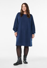 Femme portant une robe pull longue oversize bleu marine, des collants noirs et des bottines noires, se tenant contre un fond blanc.