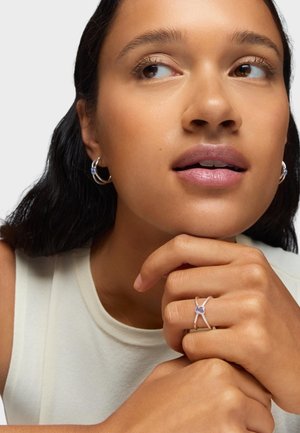 Orecchini a cerchio in argento con un accento blu e un anello in argento con una pietra viola. Top senza maniche di colore chiaro e semplice. Texture della pelle liscia.