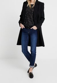 Svart kappa, prickig mörk tröja, blå jeans, uppvikta ärmar, svarta loafers. Sammanlagt en avslappnad outfit med varierande texturer och färger.