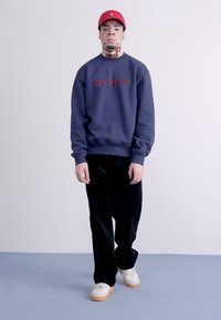 Sudadera azul marino con el logo rojo de "carhartt", combinada con pantalones de pana negra y zapatillas blancas con suelas de goma. Una gorra roja completa el conjunto.