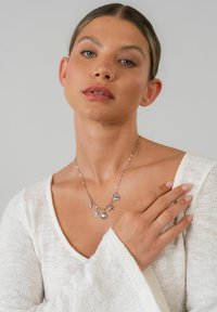 Silberne Halskette mit Charms, darunter ein Seestern, eine Schildkröte und ein runder Anhänger, getragen von einem Model in einem weißen, strukturierten Oberteil.