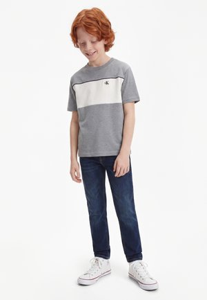 Garçon aux cheveux roux portant un t-shirt gris et blanc à manches courtes, un jean bleu foncé et des baskets blanches, regardant vers le bas et souriant.