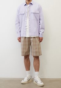Camicia button-up di un leggero viola con due tasche frontali, abbinata a pantaloni corte beige a quadri. Scarpe da ginnastica bianche con dettagli neri completano l'outfit.