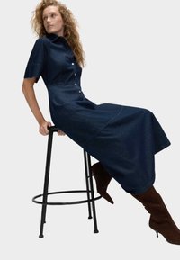 Denim skjortekjole i mørkeblå med korte ærmer, knapstolpe og en udflodet nederdel. Båret med brune knæhøje støvler.