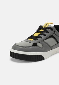 Gray leather and mesh sneaker with black outsole, yellow laces, and black accents. Features a padded collar and textured upper.