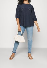 Navy blue embroidered blouse with three-quarter sleeves, light blue skinny jeans, white strappy sandals, and a cream-colored handbag.